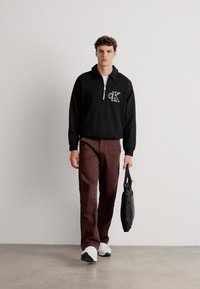 Felpa nera con zip a un quarto, con un logo "CK" grigio chiaro. Abbinata a pantaloni marroni e sneakers bianche, mentre tiene una borsa nera.