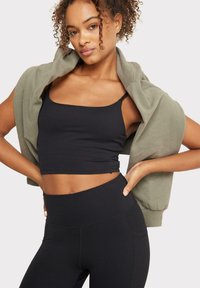 Top corto negro sin mangas y leggings de cintura alta combinados con un sudadera verde oliva colgada sobre los hombros. Tejido suave con costuras mínimas.
