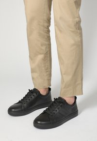Černé kožené tenisky s hladkou texturou, kulatou špičkou a šněrovacím designem. Nosené s světle béžovými kalhotami, což vytváří neformální outfit.