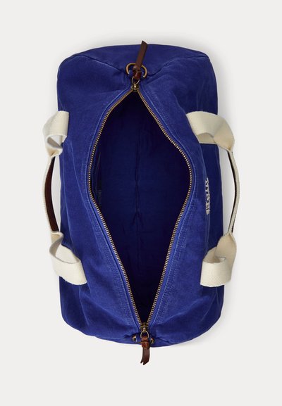 Μπλε βαμβακερή τσάντα duffel με μπεζ λαβές, ανοιχτό χρυσό φερμουάρ και δερμάτινο τράβηγμα φερμουάρ, απεικονιζόμενη από πάνω σε λευκό φόντο.