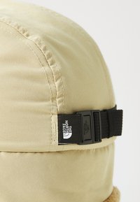 Tan bucket hat with a soft, textured brim and an adjustable black strap. Features a logo tag on the side. Made of durable fabric.