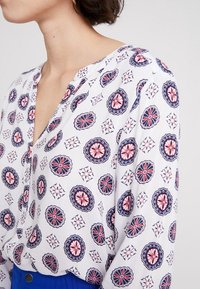 Vit blus med upprepande blommönster i röd och marinblå, med V-ringning och mjuka veck vid axlarna.