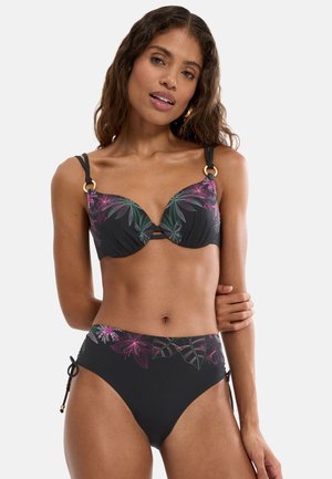 Kobieta w czarnym bikini z różowo-zielonym wzorem kwiatowym, stojąca z jedną ręką skrzyżowaną na tle neutralnym.