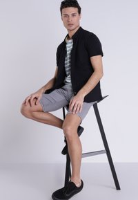 Camisa negra de manga corta sobre una camiseta de rayas, pantalones cortos gris claro y zapatillas negras, sentado en un taburete negro con un fondo neutro.