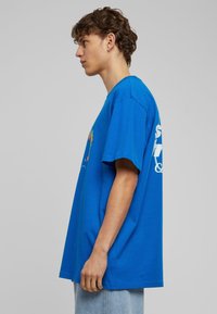 Blå bomulds t-shirt med korte ærmer, der har et farverigt grafik på forsiden og hvid tekst på bagsiden. Løs pasform.