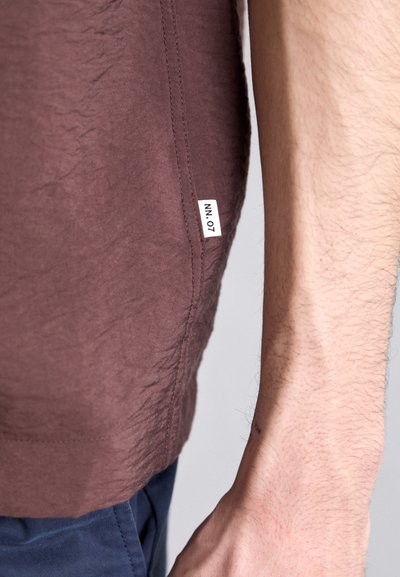 Braun, strukturiertes T-Shirt mit einem Seitenschild. Es hat ein leicht zerknittertes Design, kurze Ärmel und einen lockeren Schnitt. Getragen mit blauen Hosen.