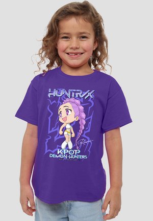 Smiling child with curly hair wearing purple "Huntrx K-Pop Demon Hunters" T-shirt featuring a cartoon character with long braided purple hair.