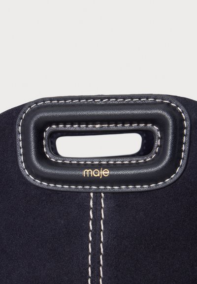 Gros plan d'une poignée de sac en cuir noir et tissu avec des coutures blanches et le logo doré "maje" sous l'ouverture de la poignée.