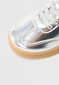 Baskets métalliques argentées avec dessus en cuir lisse, lacets plats et semelle en caoutchouc. Caractéristiques : bout arrondi et surpiqûres décoratives.