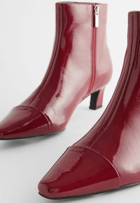 Rote Lackleder-Stiefeletten mit spitzer Zehenpartie, Reißverschluss und einem 5 cm Blockabsatz. Glatte, glänzende Textur und geschwungene Silhouette.