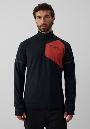 Homme aux cheveux bruns bouclés et à la barbe porte un haut noir à manches longues avec un détail de poche poitrine rouge et une demi-fermeture éclair sur un fond uni.