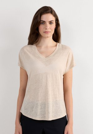 Femme aux longs cheveux bruns portant un t-shirt beige à manches courtes à col en V et un pantalon noir, debout devant un mur blanc uni.