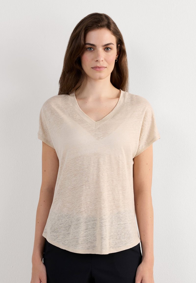 Mujer con cabello largo y castaño que lleva una camiseta beige de manga corta y escote en V y pantalones negros, de pie frente a una pared blanca lisa.