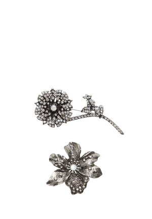 To sølvfarvede brocher: den ene prydet med et blomsterdesign dekoreret med rhinsten og en perle, mens den anden har en struktureret blomst med en perle i midten.