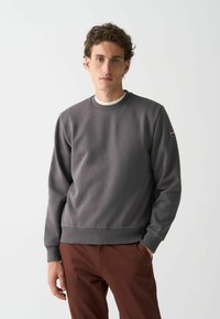 Hanorac gri dintr-un material texturat, cu guler tip crew, manșete ribbate și un patch cu logo pe mâneca stângă. Asortat cu pantaloni maronii.