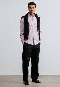 Νεαρός άνδρας στέκεται με τα χέρια στις τσέπες, φοράει ανοιχτό ροζ πουκάμισο με κουμπιά, μαύρο παντελόνι, μαύρα παπούτσια και μαύρο πουλόβερ περασμένο πάνω από τους ώμους.