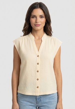 Femme aux cheveux foncés portant un chemisier beige sans manches à boutons et un jean bleu clair, face à la caméra sur un fond uni.