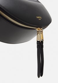 Sac en cuir noir avec des zips et des finitions dorées. Forme arrondie, logo visible sur le devant, et deux tirettes en cuir pour les zips. Texture lisse.