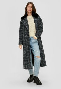 Langer schwarz-weißer karierter Mantel mit einem schwarzen Pelzkragen, kombiniert mit einem cremefarbenen Pullover und hellblauen, zerrissenen Jeans sowie schwarzen klobigen Stiefeln.