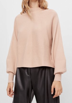 Femme portant un pull côtelé rose clair avec des manches ballon et un pantalon en simili cuir noir devant un fond uni.