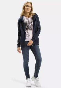 Granatowa bluza z kapturem na zamek, z ozdobnymi sznurkami, w połączeniu z jasnoróżowym t-shirtem z grafiką z roślinnymi wzorami, ciemnymi dżinsami i białymi trampkami.