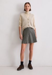 Beiges Strickweste mit Knöpfen, kombiniert mit einem hellen Rollkragenpullover. Ein grauer plissierter Rock und dunkelbraune Slipper runden das Outfit ab.