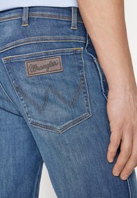 Plave traperice s vidljivom smeđom kožnom oznakom Wrangler na stražnjem džepu, naglašene kontrastnim žutim šavovima.