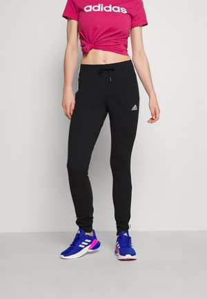 Osoba ubrana w różowy t-shirt Adidas zawiązany w talii, czarne spodnie dresowe Adidas i niebieskie buty sportowe Adidas stoi na tle jednolitego tła.