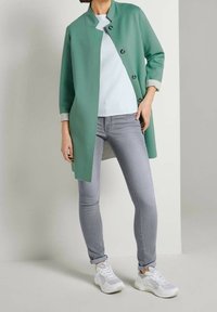 Muntgroene, knielange jas met een opstaande kraag en zwarte knopen, gecombineerd met een lichtblauw topje en grijze skinny jeans, afgewerkt met witte sneakers.