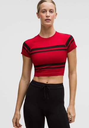 Frau trägt ein rotes, bauchfreies Sporttop mit schwarzen Streifen und schwarze Hosen mit Kordelzug, steht vor einem schlichten, hellen Hintergrund.