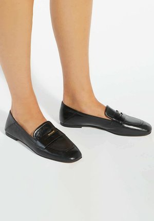 Zwarte loafers met een mix van matte en glanzende lederen texturen, puntige neus en een accentstrap met gouden logo-detail. Plat profiel.
