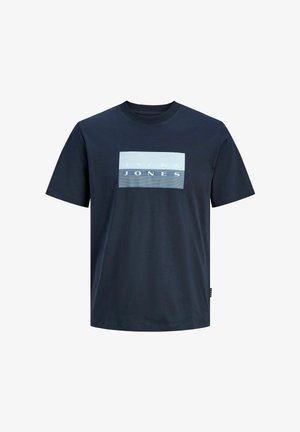 Giacca imbottita verde oliva con cappuccio, design trapuntato, abbinata a una camicia blu scuro, jeans chiari e scarpe nere.