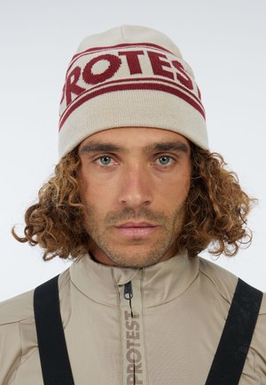 Cream knit beanie with maroon "PROTEST" text, featuring a round top. Worn by a person with curly hair, set against a neutral background.
