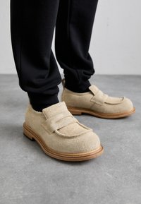 Zapatos de fieltro beige con una correa, punta redonda y suela de madera. Llevados con pantalones negros ajustados, mostrando una textura suave y un diseño minimalista.