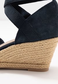 Sandale compensée en tissu noir avec des brides croisées à la cheville et un talon plateforme en jute tissé sur fond blanc.