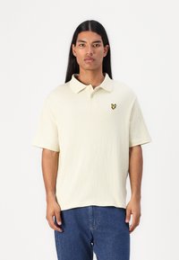 Polo à manches courtes de couleur jaune clair, en tissu texturé, avec un col et un logo brodé sur la poitrine. Porté avec un jean bleu.