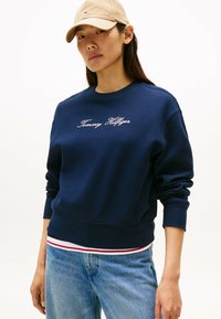 Felpa navy con polsini e orlo a coste, caratterizzata dal logo "Tommy Hilfiger" ricamato in bianco. Indossata sopra una maglietta a righe bianca e rossa.