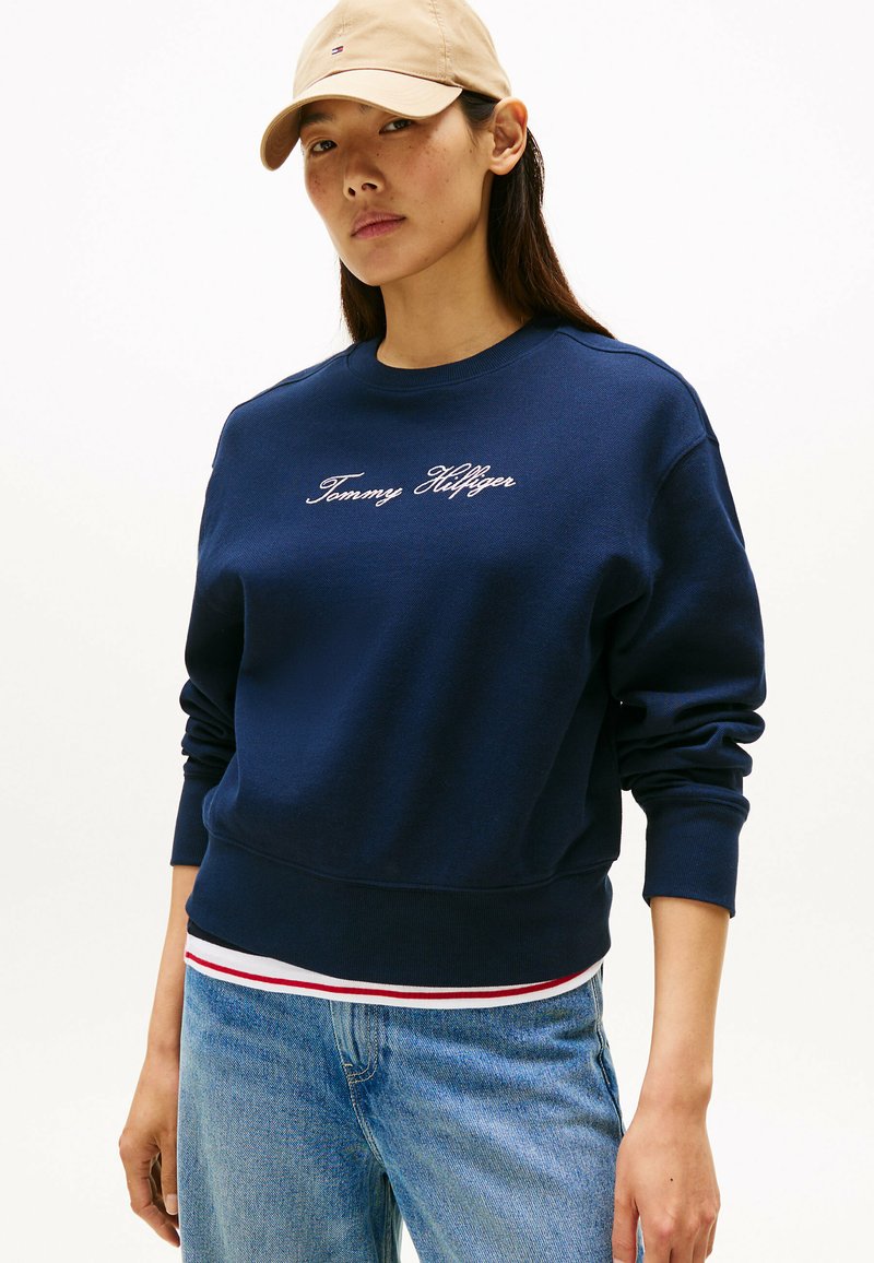 Felpa navy con polsini e orlo a coste, caratterizzata dal logo "Tommy Hilfiger" ricamato in bianco. Indossata sopra una maglietta a righe bianca e rossa.