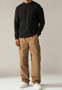 Sweat-shirt noir à manches longues associé à un pantalon cargo beige avec poches latérales, complété par des baskets blanches, sur un fond neutre.