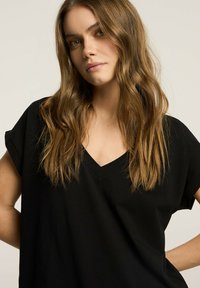 Czarna koszulka z dekoltem w serek i krótkimi, założonymi rękawami, uszyta z miękkiego materiału. Luźny krój i gładka tekstura. Model pozuje na jednolitym tle.