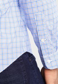 Blå och vit rutig skjorta med långa ärmar, knäppning vid handlederna och en rundad nederkant; kombinerad med mörka denimjeans.