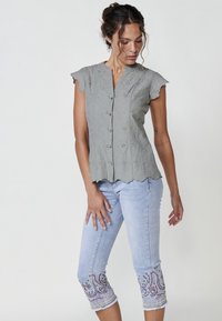 Graue, bestickte Bluse mit kurzen Ärmeln und Knopfleiste. Kombiniert mit hellblauen Jeans, die an den Säumen dekorative Stickereien aufweisen.