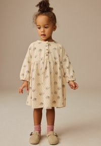 Blumengemustertes, cremefarbenes Kleid mit langen Ärmeln, Knopfdetails und gerafftem Taillengürtel; kombiniert mit rosa gerippten Socken und beigen Schuhen.