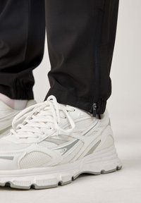 Czarne sportowe spodnie z elastycznymi mankietami, z zamkiem błyskawicznym na kostce, połączone z białymi i szarymi butami do biegania z siateczkowymi akcentami.