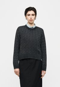 Άτομο με κοντά σκούρα μαλλιά που φοράει σκούρο γκρι πλεκτό πουλόβερ με σχέδιο καλωδίου πάνω από ανοιχτό μπλε πουκάμισο με γιακά και μαύρη φούστα σε ουδέτερο φόντο.