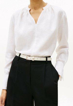 Blouse blanche à manches longues avec col froncé, rentrée dans un pantalon noir taille haute ceinturé d'une ceinture blanche à boucle dorée.