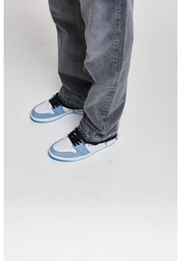 Personne portant des baskets bleu clair et blanches avec des lacets noirs et un jean gris ample, debout sur un sol blanc.