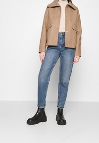 Brun uld-blandet jakke med spids krave og synlig knaplukning, parret med højtaljede blå denimjeans og sorte støvler med tyk sål.