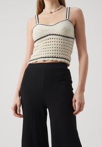Débardeur au crochet crème avec bord noir, présentant un motif texturé et des bretelles ajustables. Assorti à un pantalon large à rayures verticales noires.