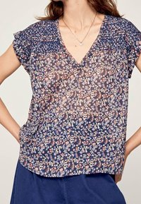 Blouse à motif floral en marine, avec un décolleté en V, des manches courtes et un empiècement smocké. Fabriquée en tissu léger et semi-transparent.